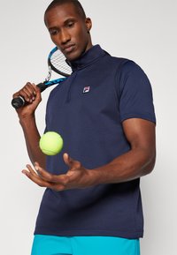 Marine teksturert polo med ståkrage og merkevarelogo, holdt av en mann. En lys grønn tennisball svever over hånden hans.