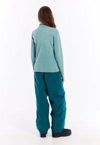 Žena otočená chrbtom, oblečená v svetlomodrom rebrovanom roláku, vo voľných tealových nohaviciach so zadnou vreckom a na čiernych platformových topánkach na bielom pozadí.