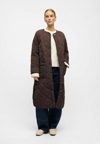 Femme portant un long manteau matelassé marron foncé sur un pull blanc, un pantalon jeans large foncé et des mocassins beige clair, se tenant devant un fond uni.