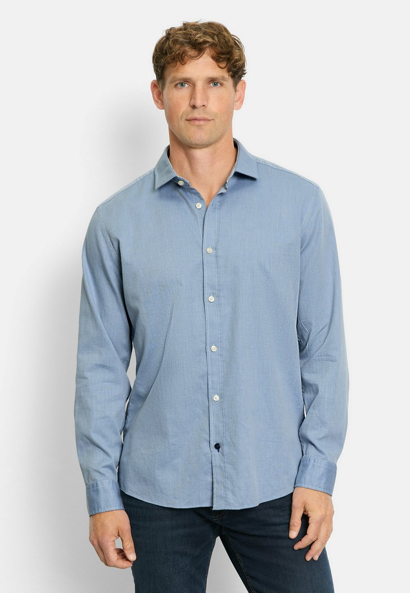Mann mit lockigem Haar, der ein hellblaues Hemd und dunkle Jeans trägt und vor einem weißen Hintergrund steht.