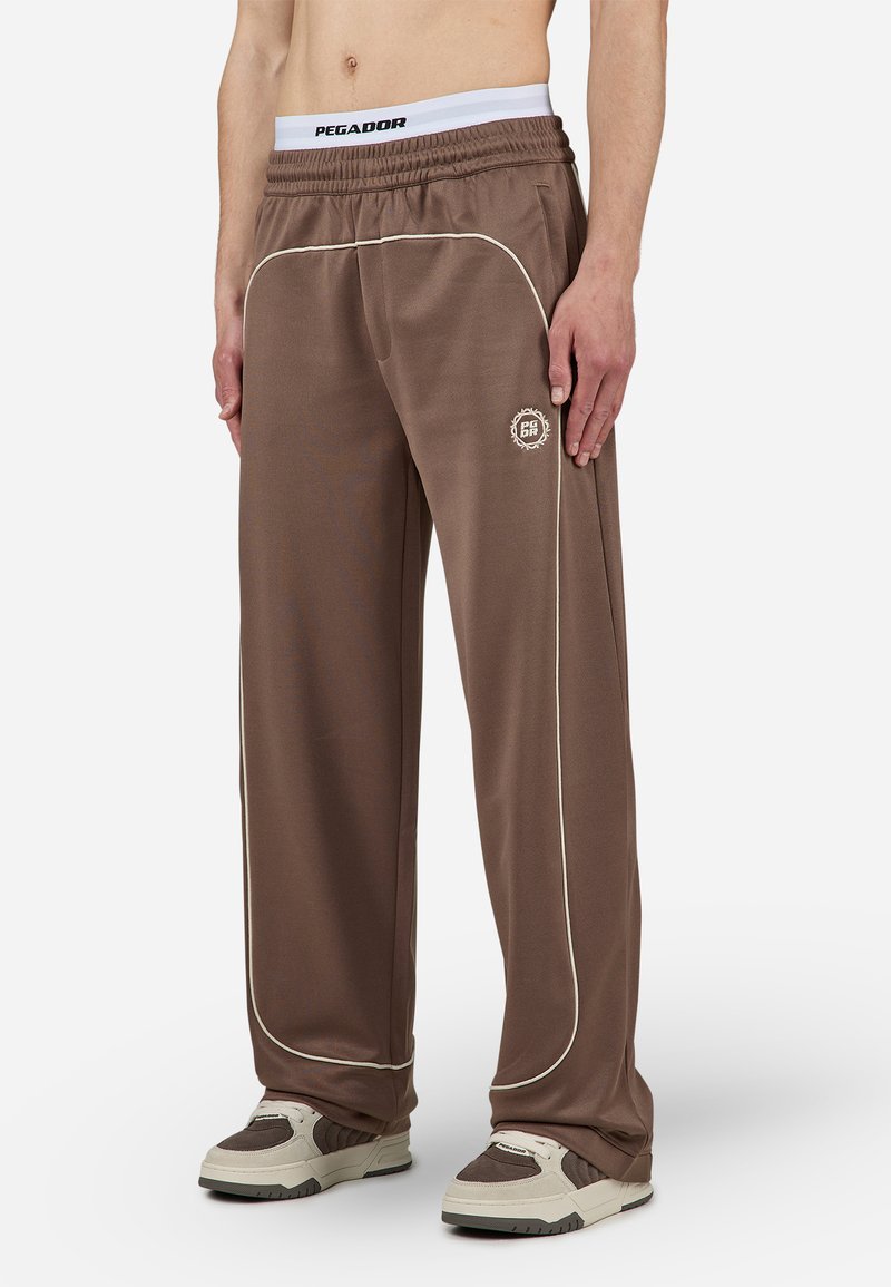 Pantalons de survêtement marron avec des passepoils blancs, taille élastique et détail de logo. Fabriqués en matériau léger et lisse. Présentent une coupe décontractée.