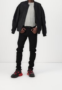 Chaqueta bomber negra, suéter gris con canalé, vaqueros negros y zapatillas negro-rojas. La chaqueta tiene un cierre frontal, un corte relajado y puños de canalé.