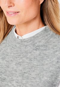Hellgrauer Strickpullover mit gerippter Textur und hohem Kragen, übereinander getragen mit einem weißen Kragenhemd, das einen Nahaufnahme der Stoffdetails zeigt.