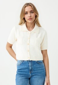 Cremfarbener kurzärmeliger Cardigan mit kragenförmigem Ausschnitt, zwei vorderen Taschen und runden Knöpfen, aus weichem, strukturiertem Material gefertigt.