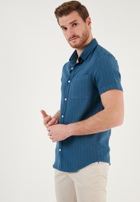 Camisa de botones de manga corta en azul oscuro, con un único bolsillo en el pecho, tela texturizada y cuello puntiagudo. Cierre de botones suave.