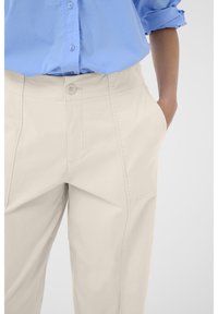 Pantalons beige clair avec une texture lisse, dotés de poches latérales en biais et d'une fermeture à bouton. Portés avec une chemise bleu à boutons.