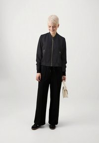 Sort cropped bomberjakke med en guldzippet lynlås og to forlommer, kombineret med løstsiddende sorte bukser og en lysefarvet håndtaske.