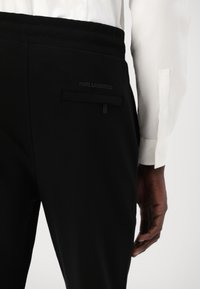 Schwarze Hosen mit glatter Textur, elastischem Bund, Seitentasche und einem dezenten Logo auf der Rückseite. Kombiniert mit einem weißen Hemd.
