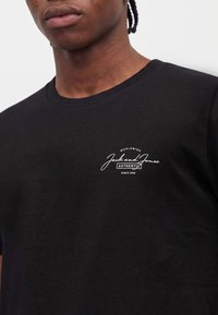 Νέος με πλεξούδες που φοράει μαύρο T-shirt με μικρό λευκό λογότυπο "Jack and Jones Authentic" στο στήθος.