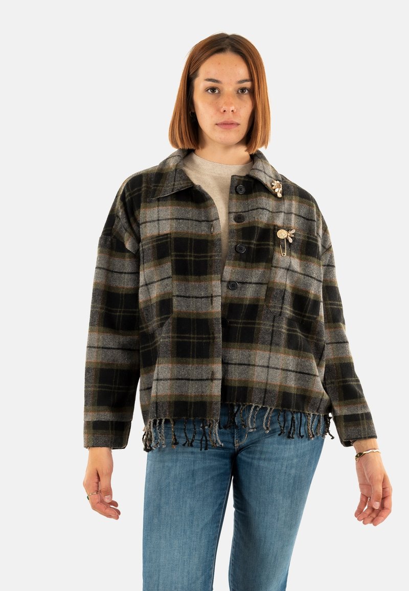Mujer con cabello lacio hasta los hombros, lleva una chaqueta de cuadros en gris y negro con flecos, un suéter beige, jeans azules y alfileres decorativos en el collar.