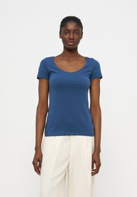 Woman with dark skin wearing a blue short-sleeve scoop neck top and white wide-leg pants, standing against a plain white background.