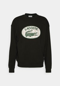 Sudadera negra con un logo de cocodrilo verde bordado en forma redonda y el texto "LACOSTE". Conta con cuello redondo y puños de canalé.