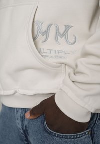 Tmavě hnědá ruka v kapsách modrých džín, oblečená v krémové mikině se stříbrně vyšitým logem "Multiply Apparel" blízko kapsy.