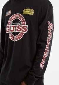 Zwarte lange mouwen T-shirt met een groot vintage-stijl "GUESS" logo, gele "GUESS Originals" patch en een afbeelding van de Amerikaanse vlag op de borst.