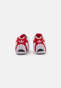 Pantofi sport albi cu accente roșii, având o suprafață texturată, guler căptușit și un logo pe călcâi. Talpă din cauciuc pentru aderență.