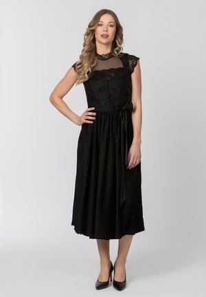 Frau steht mit der Hand in der Hüfte, trägt ein schwarzes Midikleid mit Spitzenoberteil und transparentem Ausschnitt, kombiniert mit schwarzen High Heels.