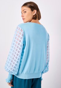 Hellblauer Pullover mit solidem Strickkörper und transparenten Ärmeln im Karomuster. Mit gerippten Bündchen und Saum, die eine lockere Passform bieten.