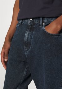 Närbild av person som bär mörkblå denimjeans med en hand i fickan och en marinblå skjorta instoppad.