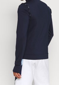 Marinblå idrottsjacka med hög krage, långa ärmar, strukturerat tyg och kontrasterande ljusblå detaljer på sidorna. Har logotypdetaljer på axlarna.