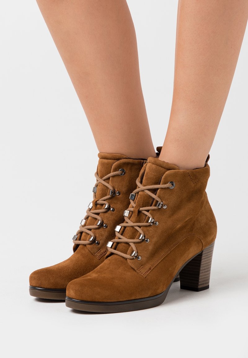 Bottines marron en daim à talons carrés avec lacets à l’avant, portées par une personne, présentées sur un fond clair uni.