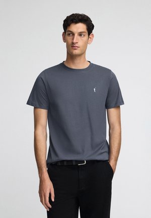 T-shirt grigio in cotone con collo rotondo e maniche corte, con un piccolo logo bianco sul petto. Abbinato a pantaloni neri.