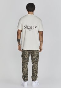 Krémové bavlněné tričko s černou grafikou "SIKSILK" na zádech, v kombinaci s maskáčovými cargo kalhotami a bílými teniskami. Jednoduchý, volný střih.