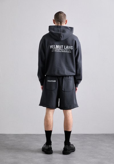 Mørkegrå Helmut Lang hettegenser med hvit tekst på baksiden, kombinert med matchende shorts med rå kant og lommer. Svart sko og sokker.