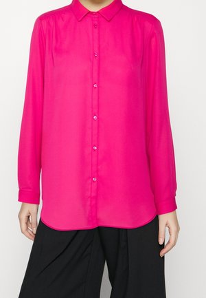 Personne portant un chemisier boutonné à manches longues rose vif, associé à un pantalon noir ample, montré du cou jusqu’aux hanches.