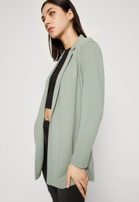 Light green blazer with a relaxed fit, long sleeves, and no buttons. Features a shawl collar and smooth fabric texture. Worn over a black top.