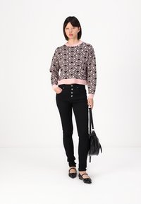 Häkelpullover mit pinkem und schwarzem Herzmuster, kombiniert mit hoch taillierten schwarzen Jeans und schwarzen Ballerinas. Das Model hält eine schwarze Handtasche.