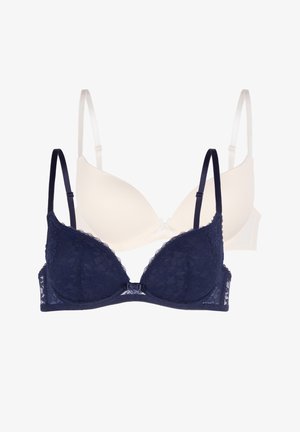 Femeie blondă zâmbitoare, purtând un set asortat de sutien și chiloți din dantelă albastru închis, stând în fața unui fundal gri simplu.