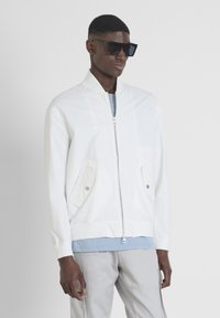 Cazadora bomber blanca con cierre frontal, bolsillos laterales y puños de canalé. Llevada sobre una camisa azul clara, combinada con pantalones grises y gafas de sol negras.