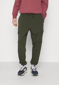 Olijfgroene cargo shorts met elastische tailleband en trekkoord, met zijzakken. Gecombineerd met marineblauwe en witte sneakers en een kastanjebruine sweatshirt.