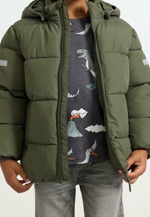 Kind trägt eine offen stehende olivgrüne Puffjacke über einem dunkelgrauen T-Shirt mit Dinosauriern, Vulkanen, Wolken und grauen Jeans.