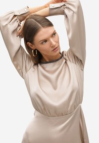 Vestido de satén beige con mangas largas abullonadas y una cintura fruncida. Presenta un escote redondo adornado con un ribete de cuentas oscuras y acentos de botones.