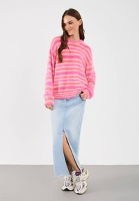 Γυναίκα που φοράει ένα ροζ φουντωτό ριγέ πουλόβερ, ελαφριά μπλε τζιν φούστα με εμπρόσθιο σκίσιμο και λευκά-μωβ αθλητικά παπούτσια, στέκεται με το χέρι στην τσέπη.