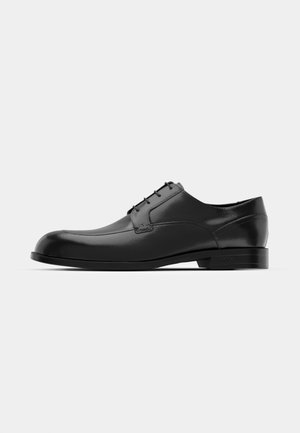 Black leather dress shoe featuring a rounded toe, smooth texture, minimal seams, and a flat sole with subtle branding on the heel.
