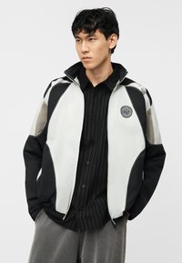 Νεαρός άντρας που φοράει μαύρο και άσπρο τζάκετ με φερμουάρ και λογότυπο στο στήθος, μαύρο πουκάμισο με λεπτές ρίγες και γκρι παντελόνι, με τα χέρια στις τσέπες, κοιτάζει αριστερά.