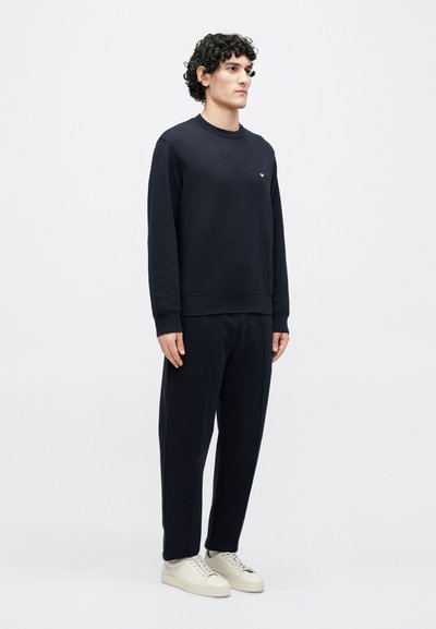 Tmavomodrá mikina s rebrovanými manžetami a spodným lemom, doplnená o zodpovedajúce nohavice. Má malú logo na hrudi a biele tenisky.