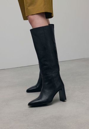 Bottes en cuir noir montantes avec un bout pointu et un talon épais, présentant une texture lisse et des détails de couture minimalistes.