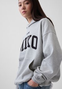 Νέα γυναίκα με μακριά σκούρα μαλλιά φοράει γκρι φούτερ με κουκούλα και το κείμενο «AIKO», με το ένα χέρι στη τσέπη, μπροστά σε απλό φωτεινό φόντο.