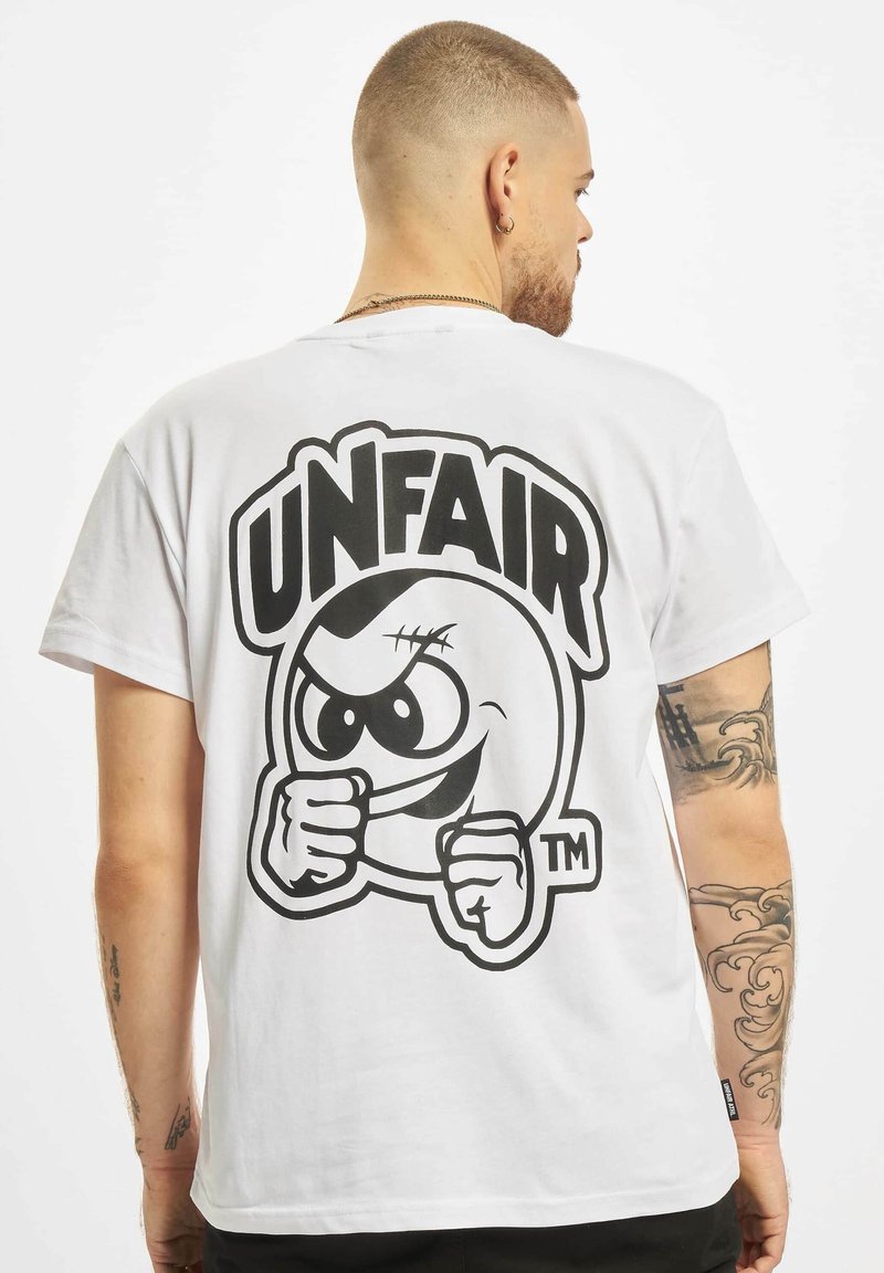 Una camiseta blanca de algodón presenta un gran gráfico negro de una cara caricaturesca con la palabra "INJUSTO" arriba, gráfico contorneado en negro.