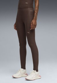 Καφέ αθλητικό κολάν με εφαρμοστή σχεδίαση, με ψηλή μέση και λεπτομέρειες με λογότυπα, συνδυασμένο με λευκά και ροζ αθλητικά παπούτσια.