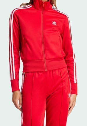 Femme portant un survêtement rouge Adidas avec des bandes blanches sur les manches et les pantalons, et des poches zippées sur la veste et le pantalon.