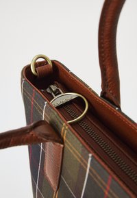 Καφέ τσάντα με δερμάτινη λαβή και καρό ύφασμα σε γήινα χρώματα. Διαθέτει φερμουάρ με μεταλλικό τράβηγμα και καφέ δερμάτινη λεπτομέρεια κοντά στη λαβή.