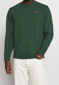 Uomo che indossa un sudore a maniche lunghe Levi's verde scuro e pantaloni bianchi, con la mano in tasca e un braccialetto d'argento al polso.