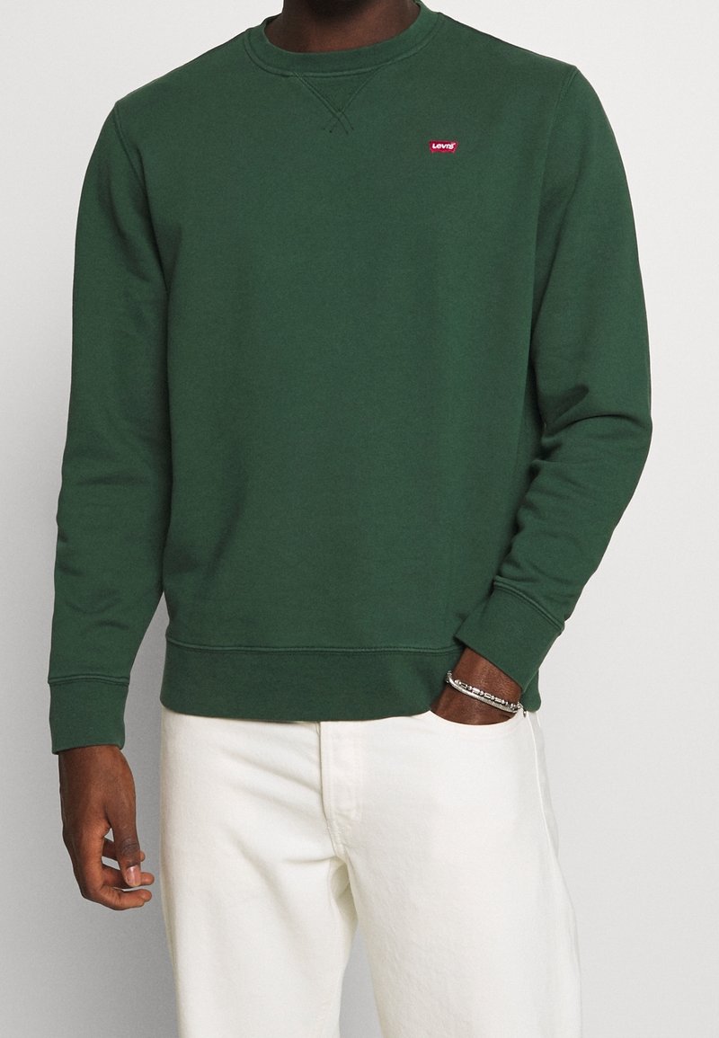 Uomo che indossa un sudore a maniche lunghe Levi's verde scuro e pantaloni bianchi, con la mano in tasca e un braccialetto d'argento al polso.