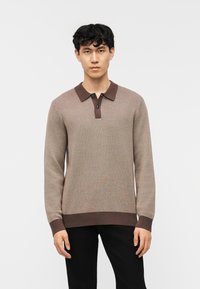 Νέος άνδρας που φοράει καφέ μακρυμάνικο πουλόβερ πόλο με σκούρο γιακά και μανσέτες, συνδυασμένο με μαύρο παντελόνι, στέκεται μπροστά σε λευκό φόντο.