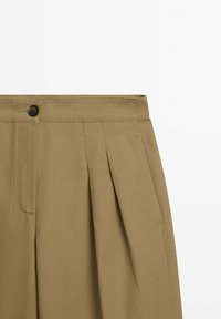 Pantalon kaki taille haute, devant plissé, fermeture par un bouton noir unique et poches latérales. Texture de tissu lisse et léger.