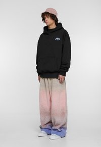 Sudadera negra con capucha y bolsillo frontal. Pantalones de chándal holgados y multicolores con un degradado de rosa a azul y zapatillas blancas. Cabello rizado y gorra rosa.